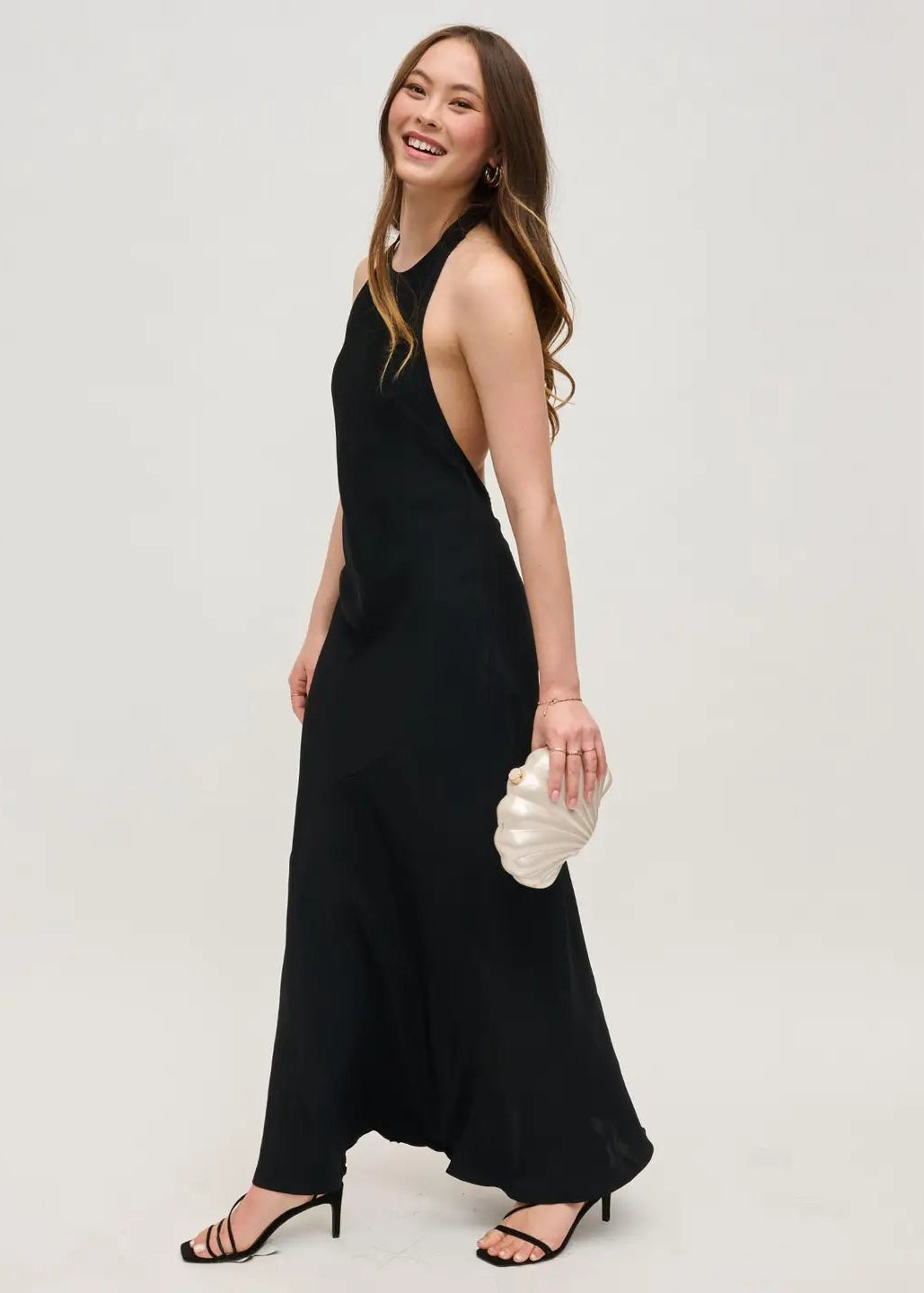 Woman wearing a black sleeveless dress holding a white clutch against a plain background