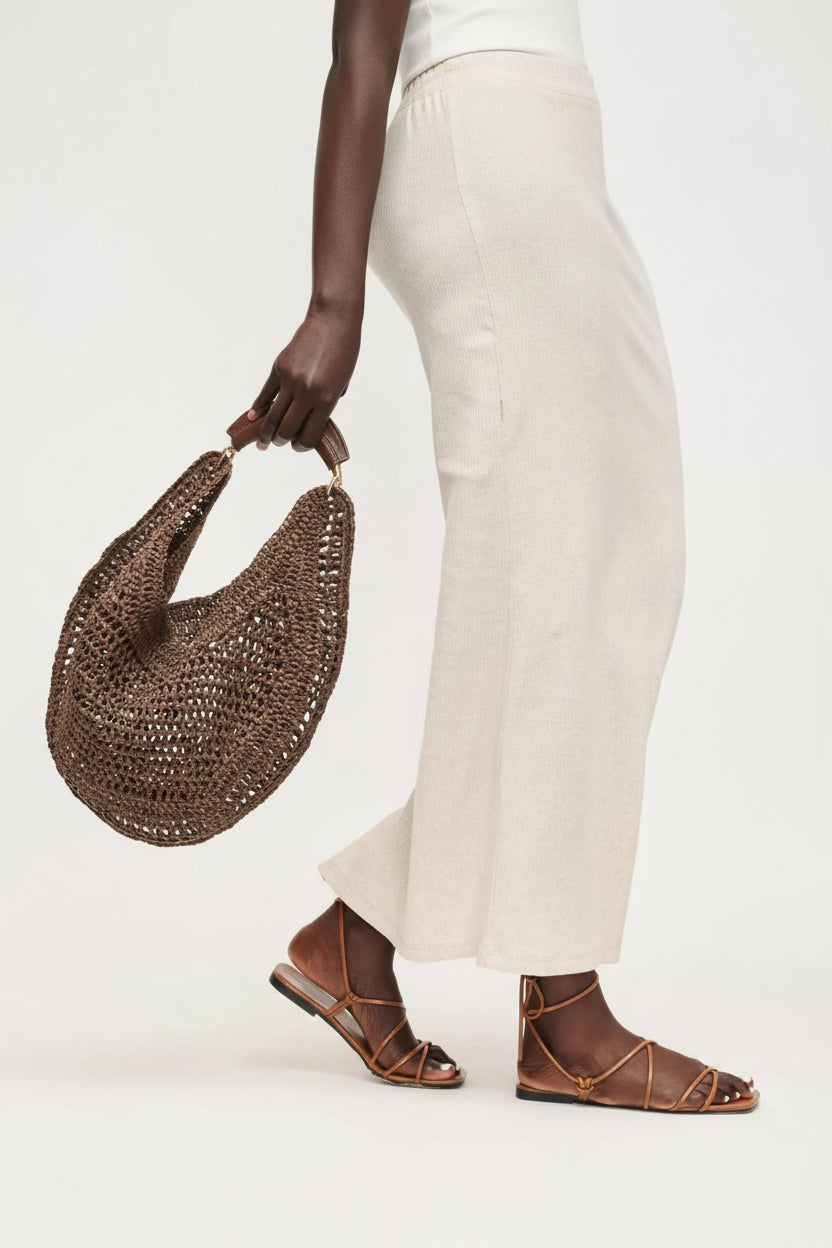 Person holding a woven brown handbag with white pants and brown sandals on a light background