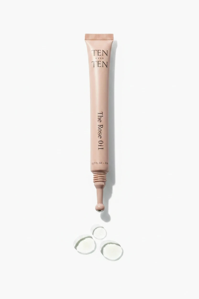 A tube of Ten Over Ten - The Rose Oil with a metallic cap, shown alongside a few drops of the oil outside the tube.