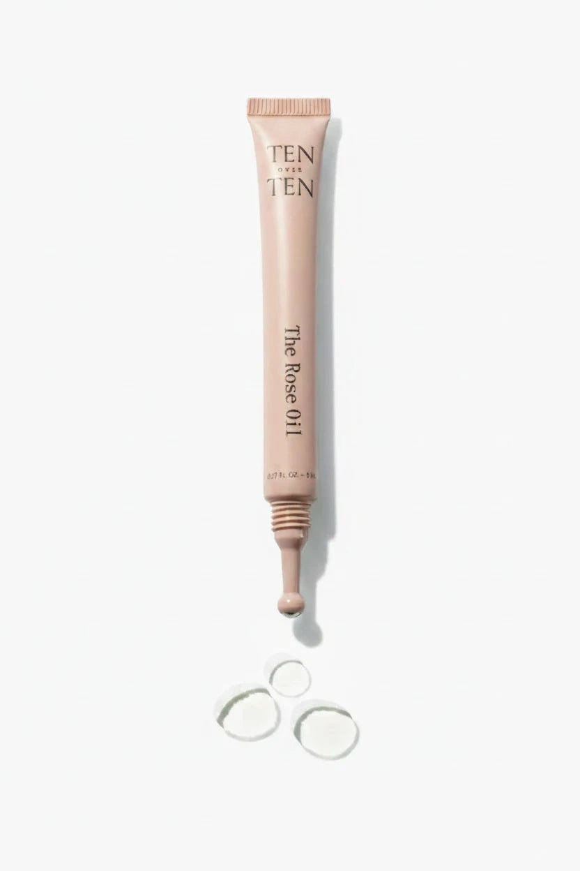A tube of Ten Over Ten - The Rose Oil with a metallic cap, shown alongside a few drops of the oil outside the tube.