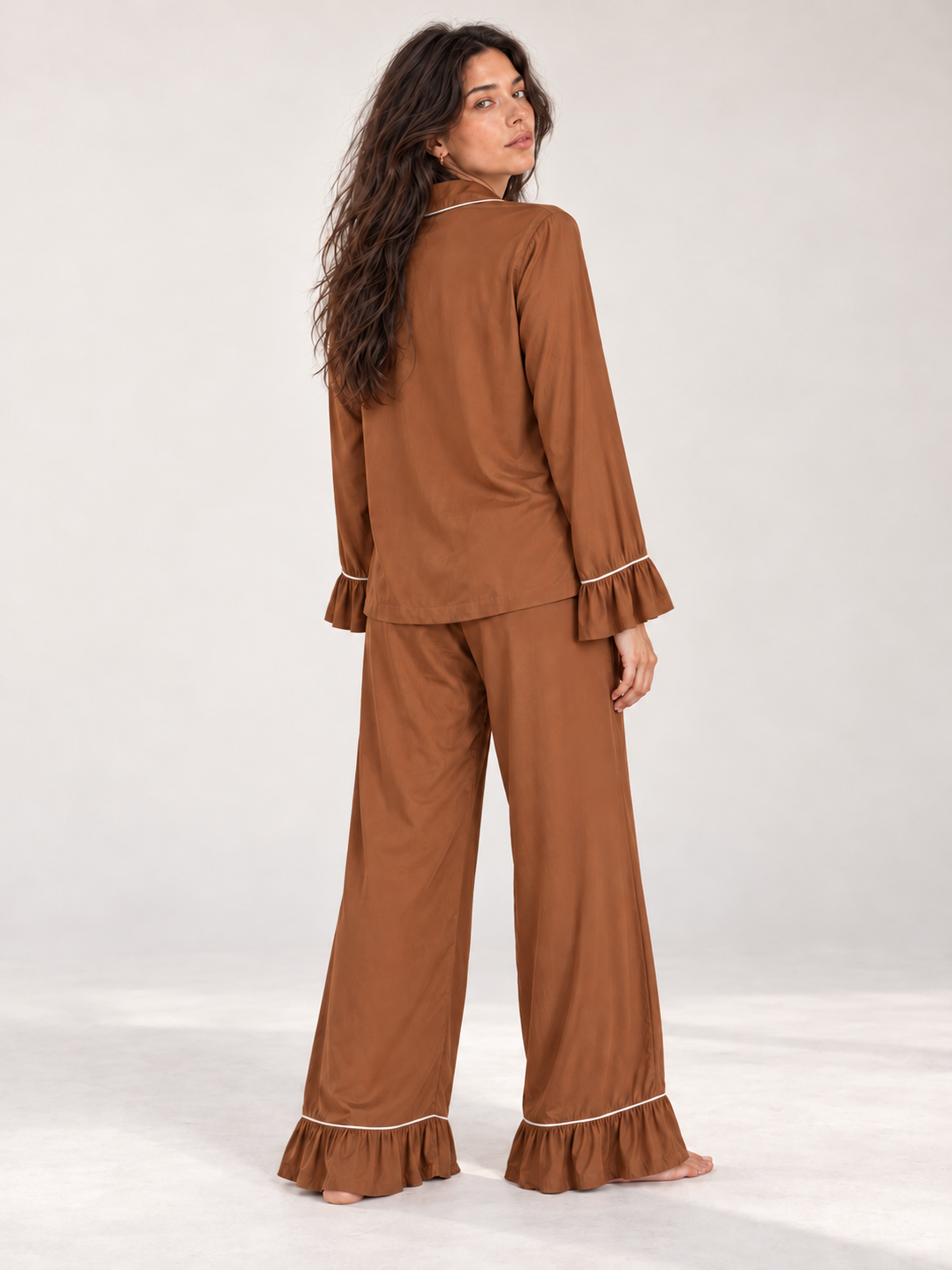 A woman modeling a two-piece pajama set in a warm brown color, shown from the back. The set includes a long-sleeve top with ruffled cuffs and matching wide-leg pants with ruffled hems, both featuring thin white piping details.