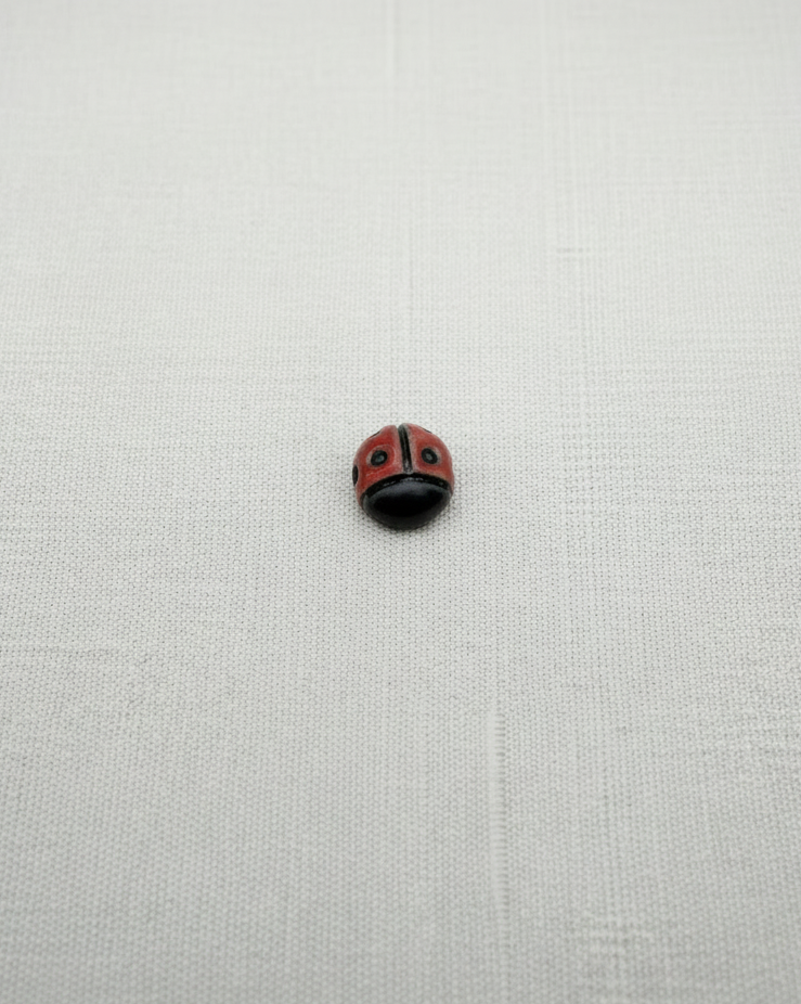 A small round ladybug bead, featuring a red and black painted body with white accents, displayed on a textured off-white fabric background.