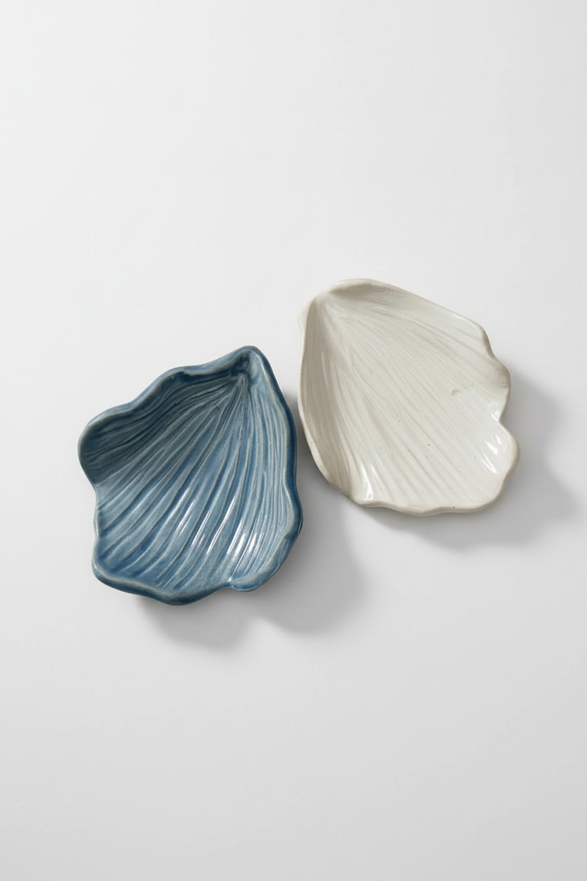 Two ceramic shell-shaped plates on a white textured background, one in blue and one in white.