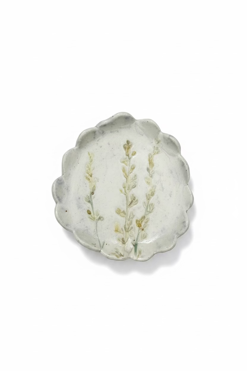 A scalloped oval ceramic plate or dish featuring a hand-painted botanical design of three light green and beige flower stems on a textured off-white background, displayed on a white linen surface.