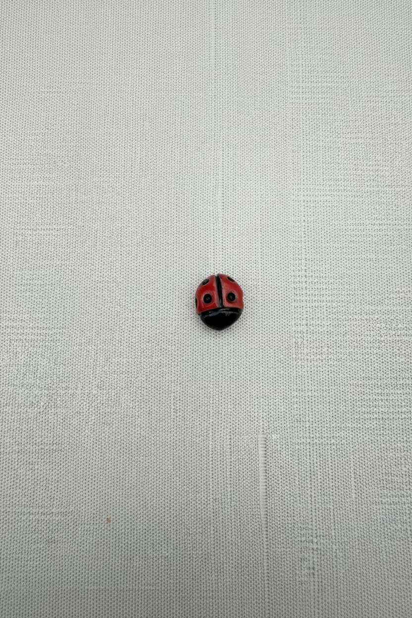 A small round ladybug bead, featuring a red and black painted body with white accents, displayed on a textured off-white fabric background.
