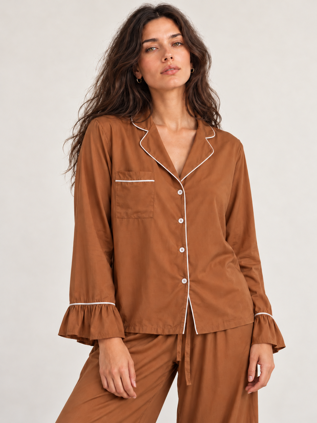 A woman modeling a two-piece pajama set in a warm brown color with white piping details, consisting of a long-sleeve button-up top with ruffled cuffs and matching drawstring pants, shot against a light gray studio background.