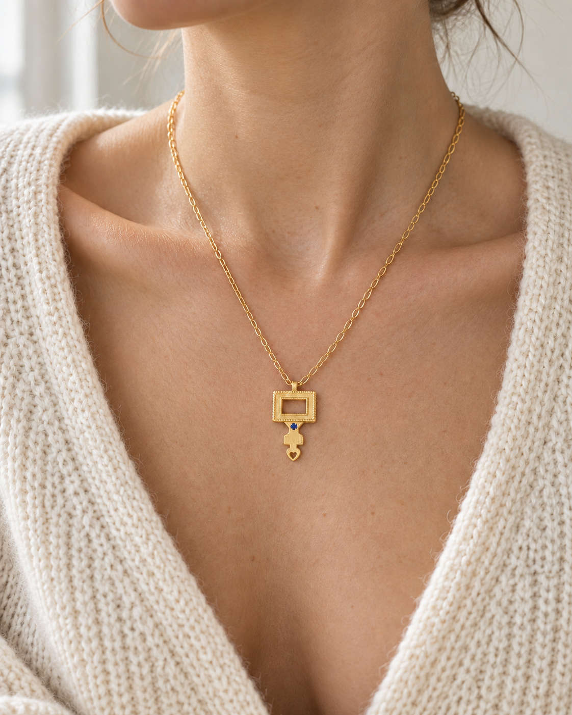 A close-up of a model wearing a delicate gold chain necklace with a geometric pendant and a small dangling charm, styled with a cream knit sweater.
