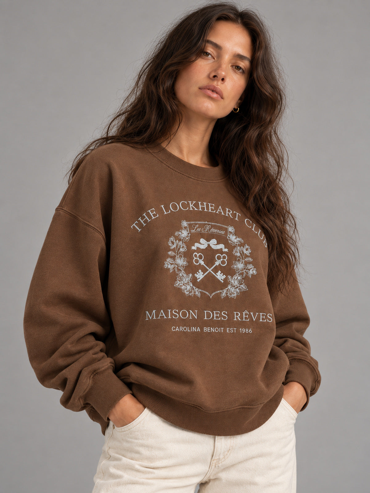 A woman wearing a brown crewneck sweatshirt with a white graphic print that reads 'THE LOCKHEART CLUB' and 'MAISON DES RÈVES', paired with white pants, against a solid gray studio background.