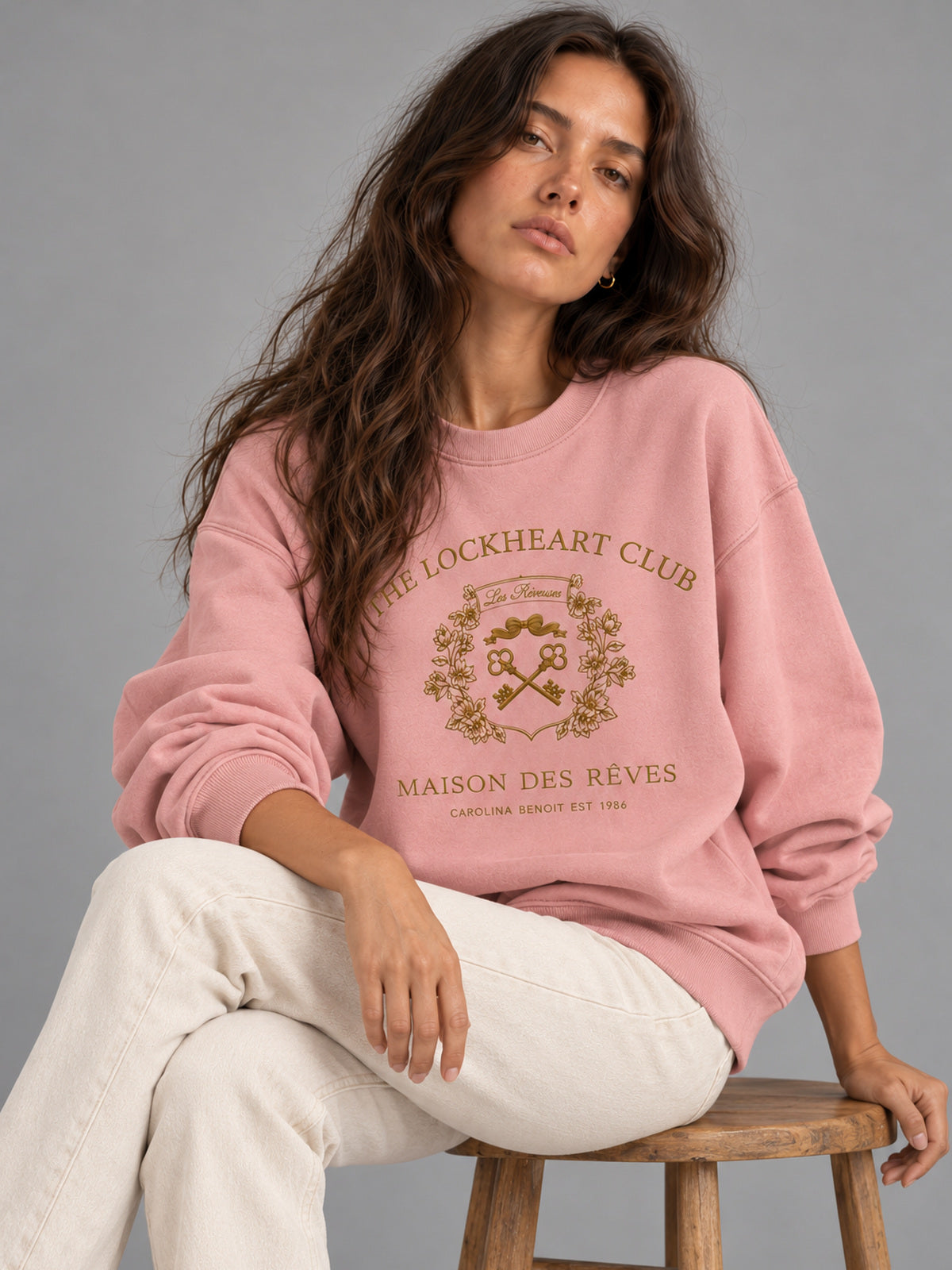 A woman sits on a wooden stool wearing a dusty pink crewneck sweatshirt with a gold-colored 'The Lockheart Club' graphic on the front, paired with off-white pants, against a solid gray studio background.