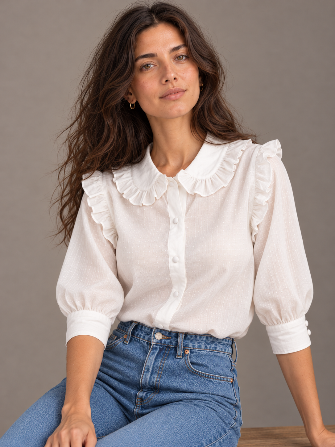 A woman wearing a white long-sleeve blouse with a large ruffled Peter Pan collar and puff sleeves, paired with blue jeans, posed against a neutral gray studio background.