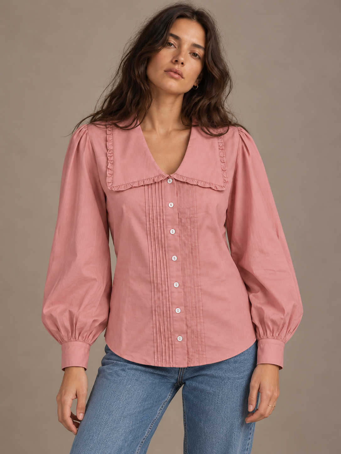 A woman wearing a dusty pink long-sleeve blouse with a ruffled collar and front buttons, paired with blue jeans, against a plain taupe background.