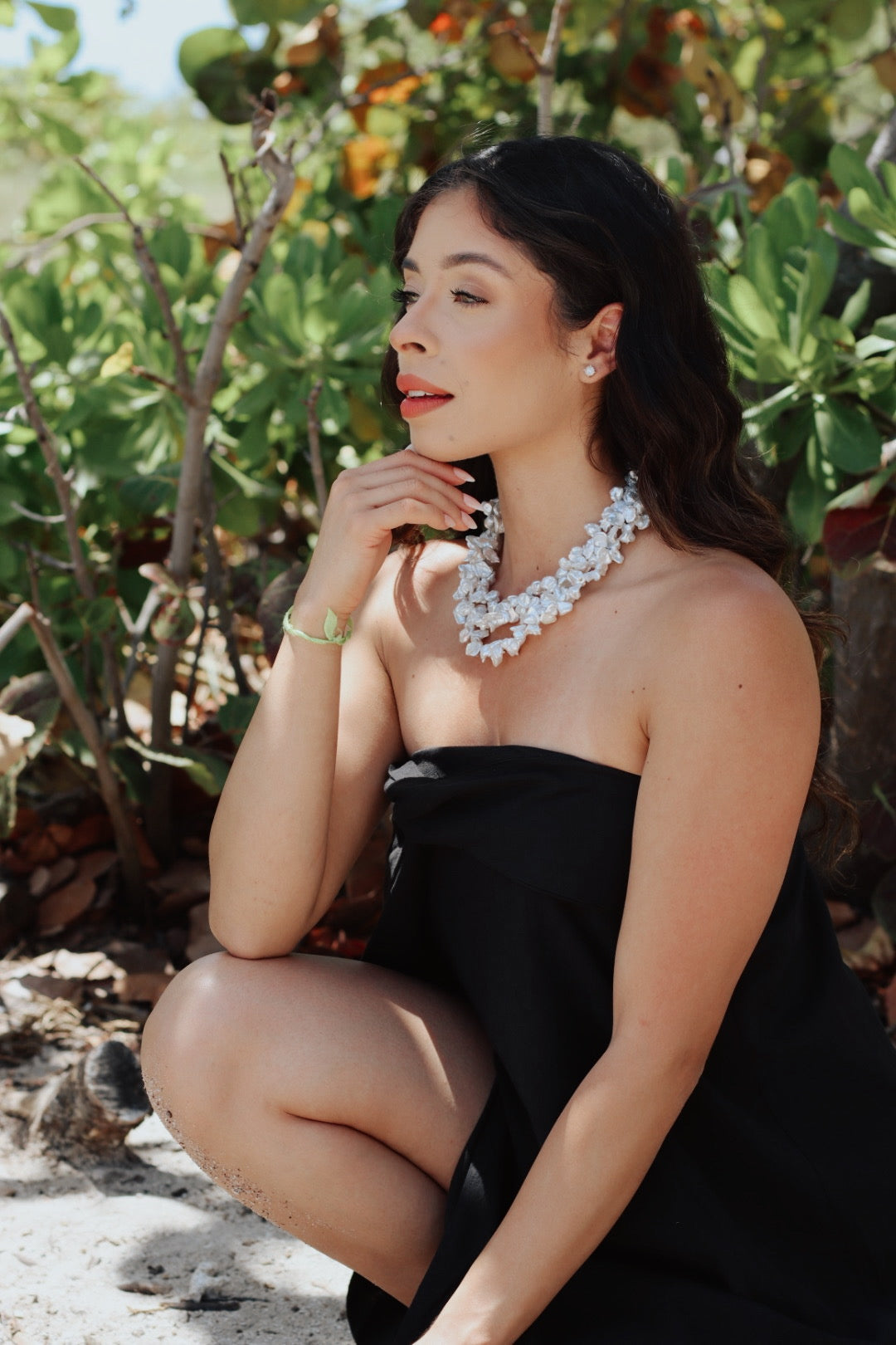 Side view of woman wearing the Baroque Garden Necklace, showing the sculptural pearls layered around her neck, seated among tropical greenery