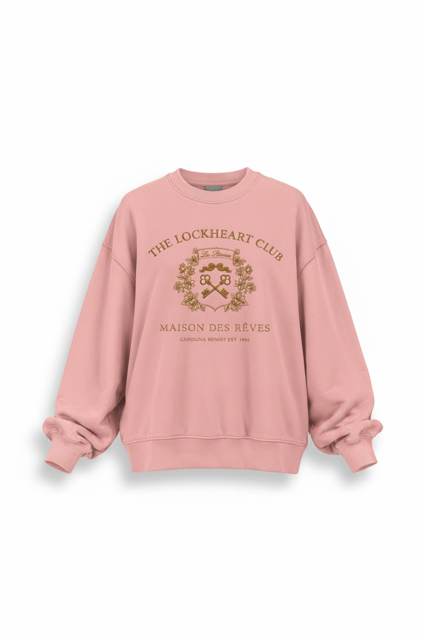 A light pink crewneck sweatshirt with a gold graphic print on the front reading 'THE LOCKHEART CLUB' and 'MAISON DES RÈVES', displayed against a light off-white background.