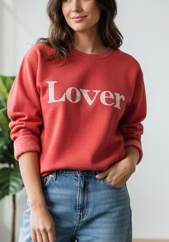 Woman wearing a red sweater with 'Lover' text and blue jeans in a bright room.