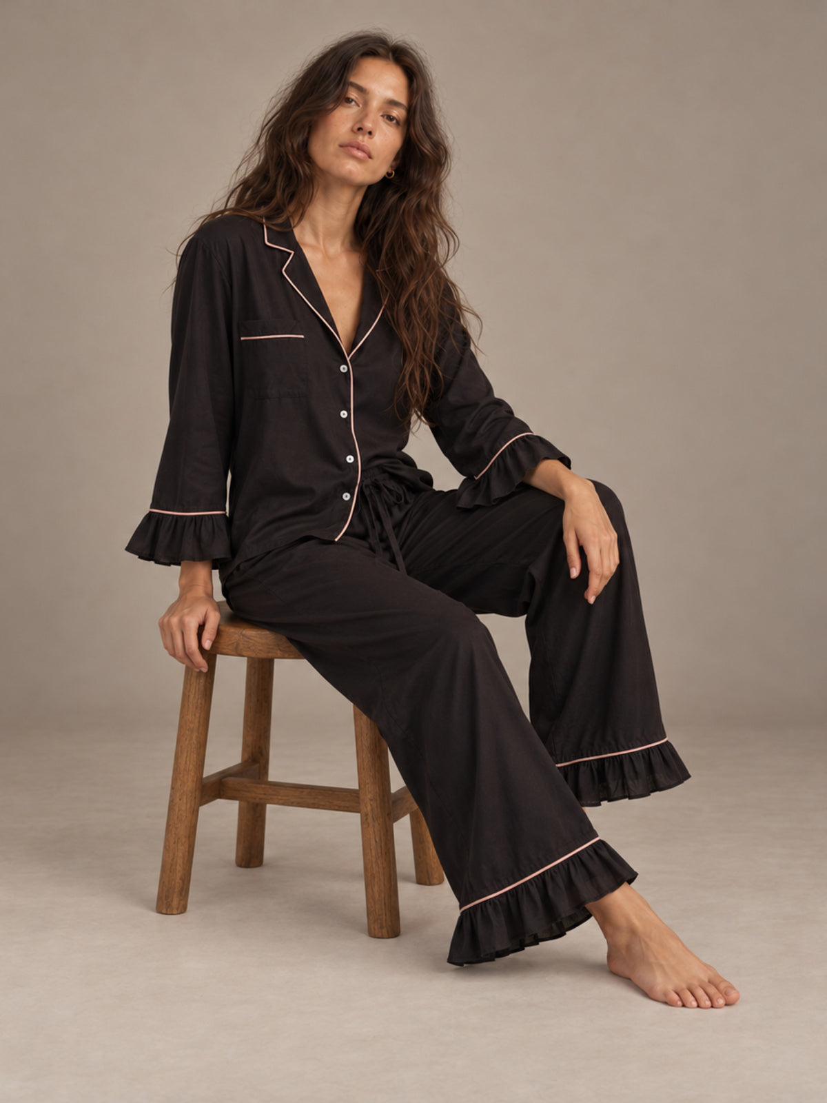 A woman wearing a black pajama set with light pink piping sits on a wooden stool against a neutral beige studio background.