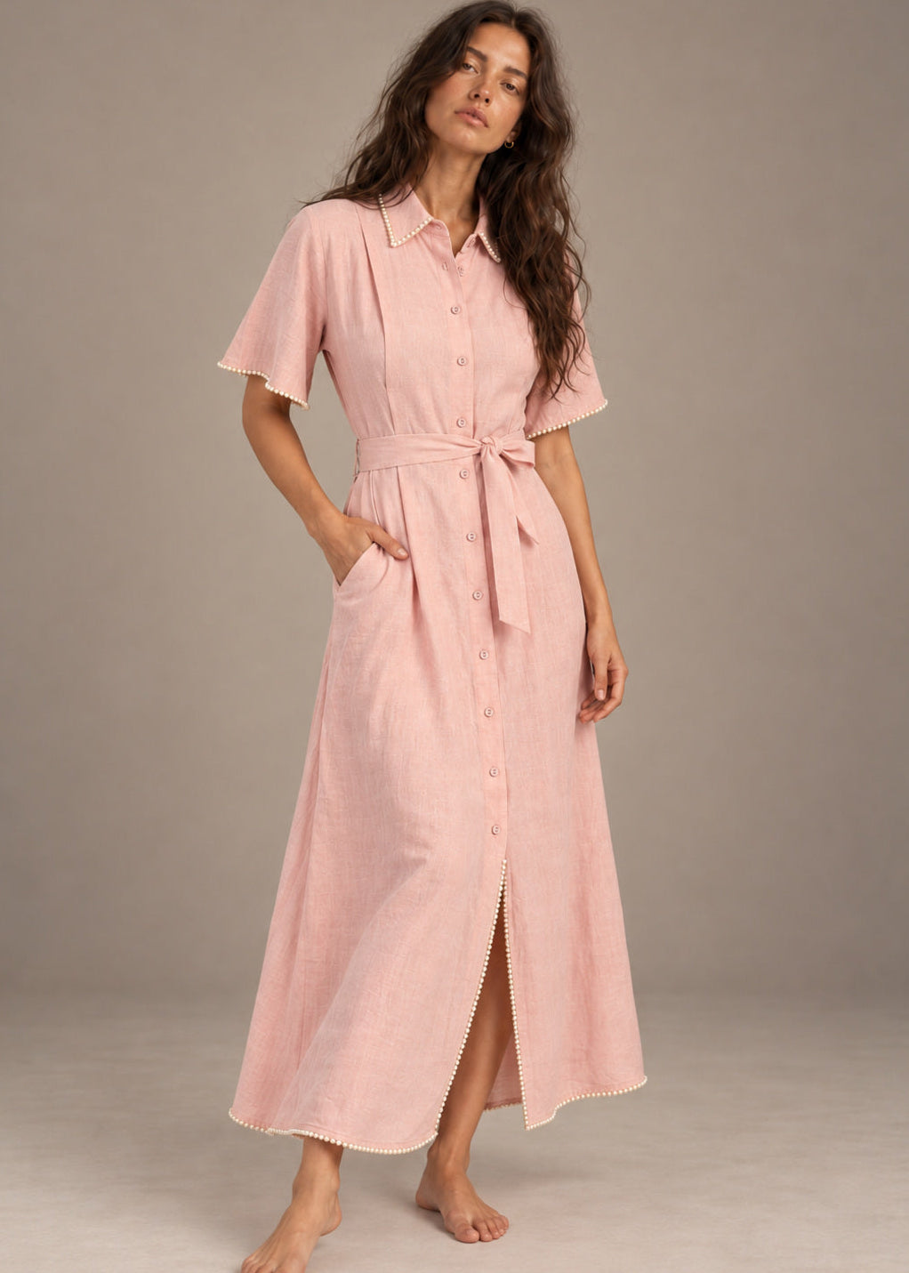 A woman modeling a light pink short-sleeve midi shirt dress with a tie waist belt, standing barefoot against a neutral studio backdrop.