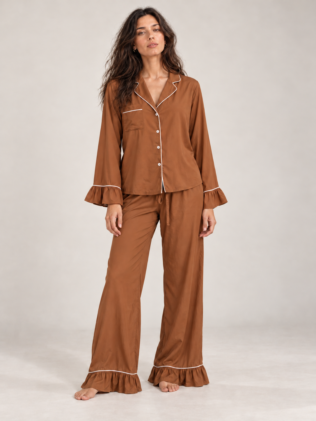 A woman modeling a two-piece pajama set in a warm brown color, featuring a long-sleeve button-up top with ruffled cuffs and matching wide-leg pants with ruffled hems, both accented with white piping.