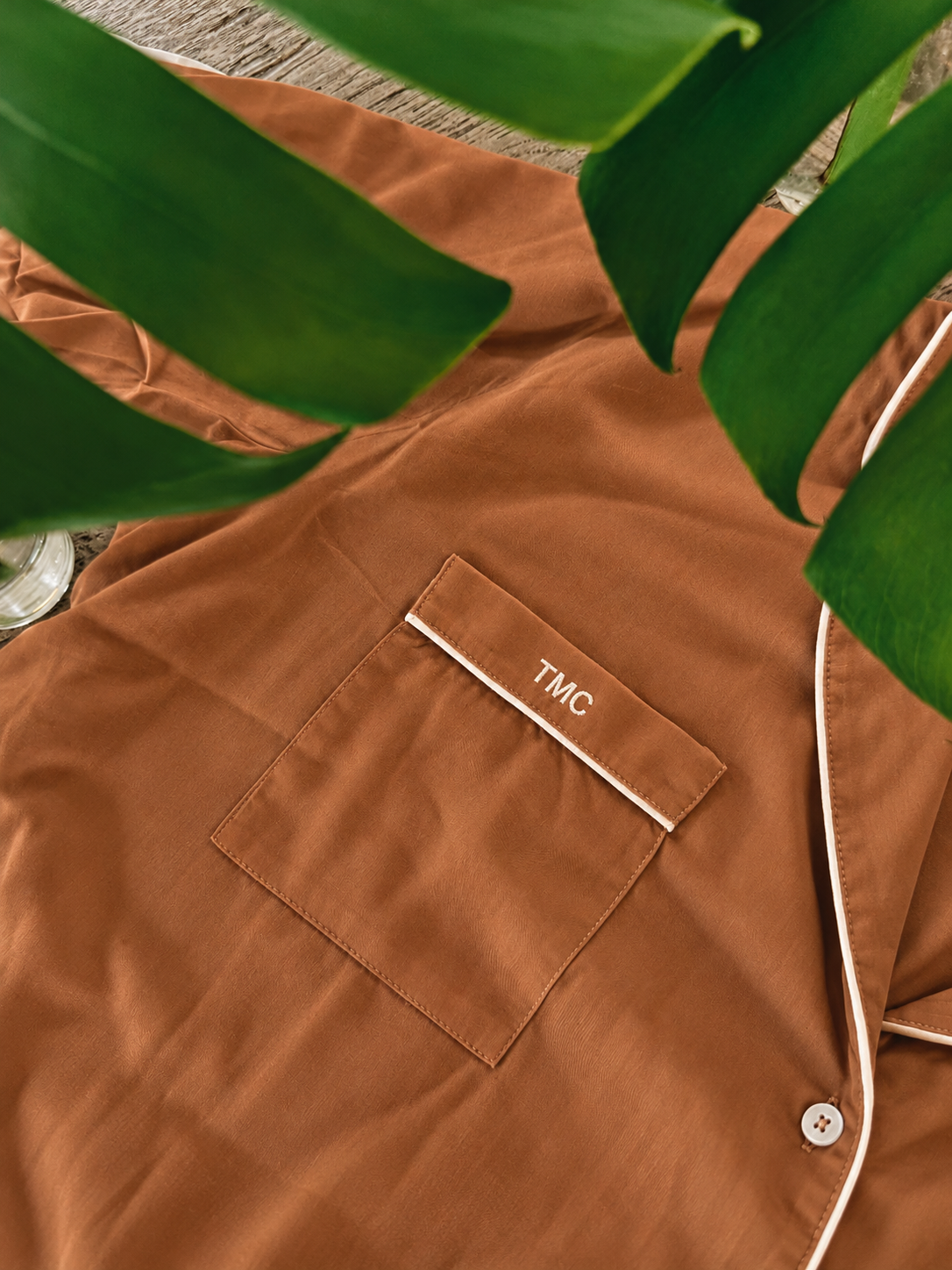 A close-up of a brown pajama shirt with white piping and a chest pocket embroidered with 'TMC'. Large green tropical leaves frame the top of the image.