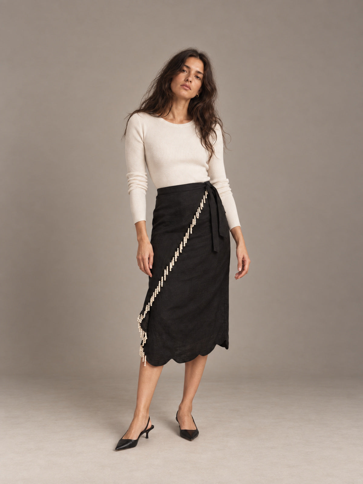 A woman modeling a black wrap midi skirt with a cream-colored braided trim detail, paired with a cream long-sleeve top and black pointed-toe heels, against a neutral gray studio background.