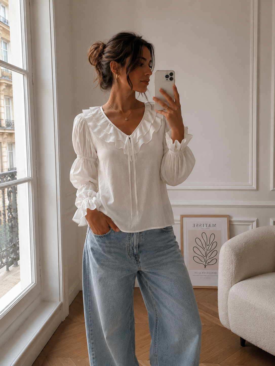 Woman taking a mirror selfie wearing a white blouse and blue jeans in a room with a window and armchair.