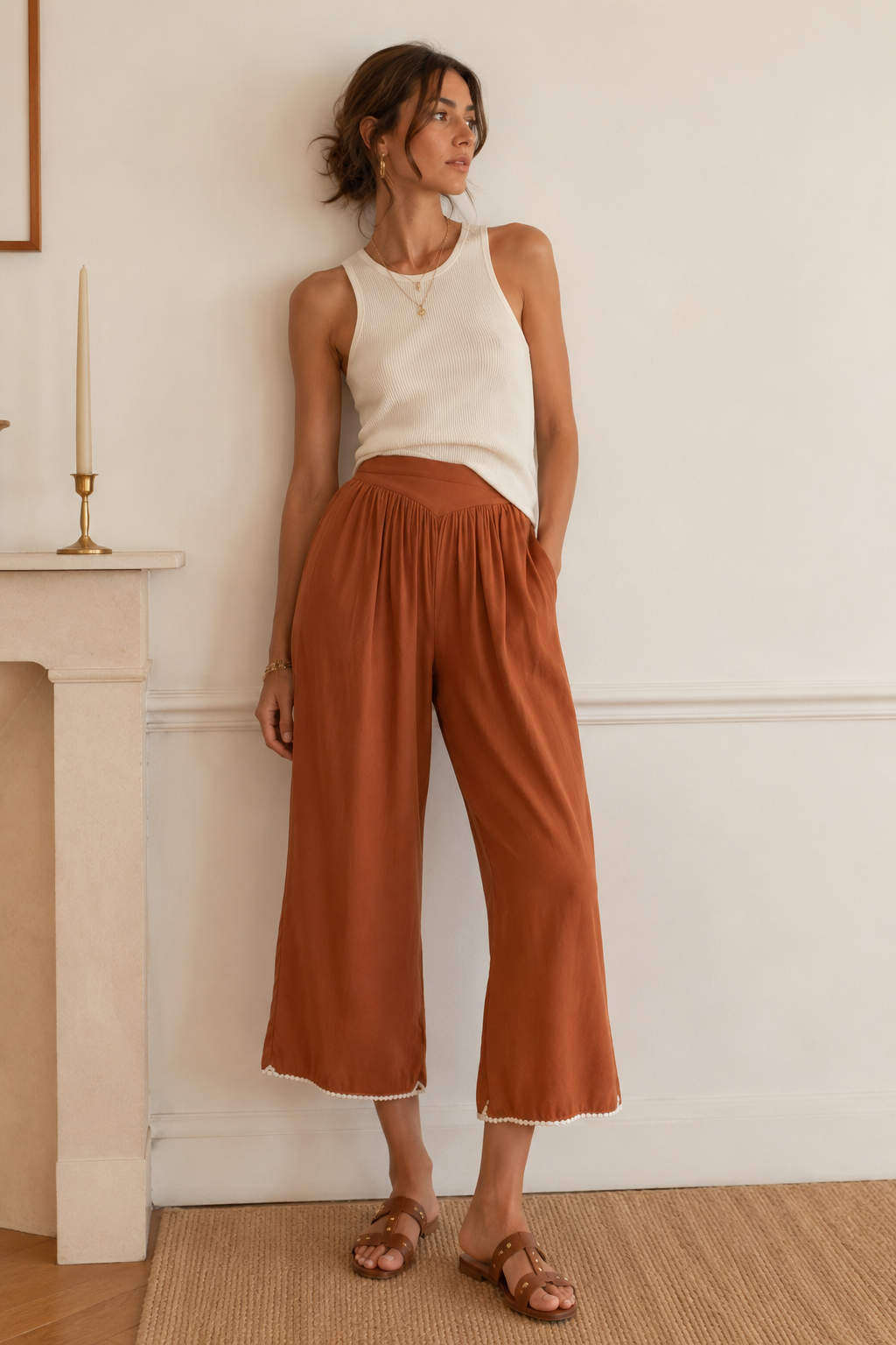 Woman wearing a white sleeveless top and brown wide-leg pants standing against a white wall.