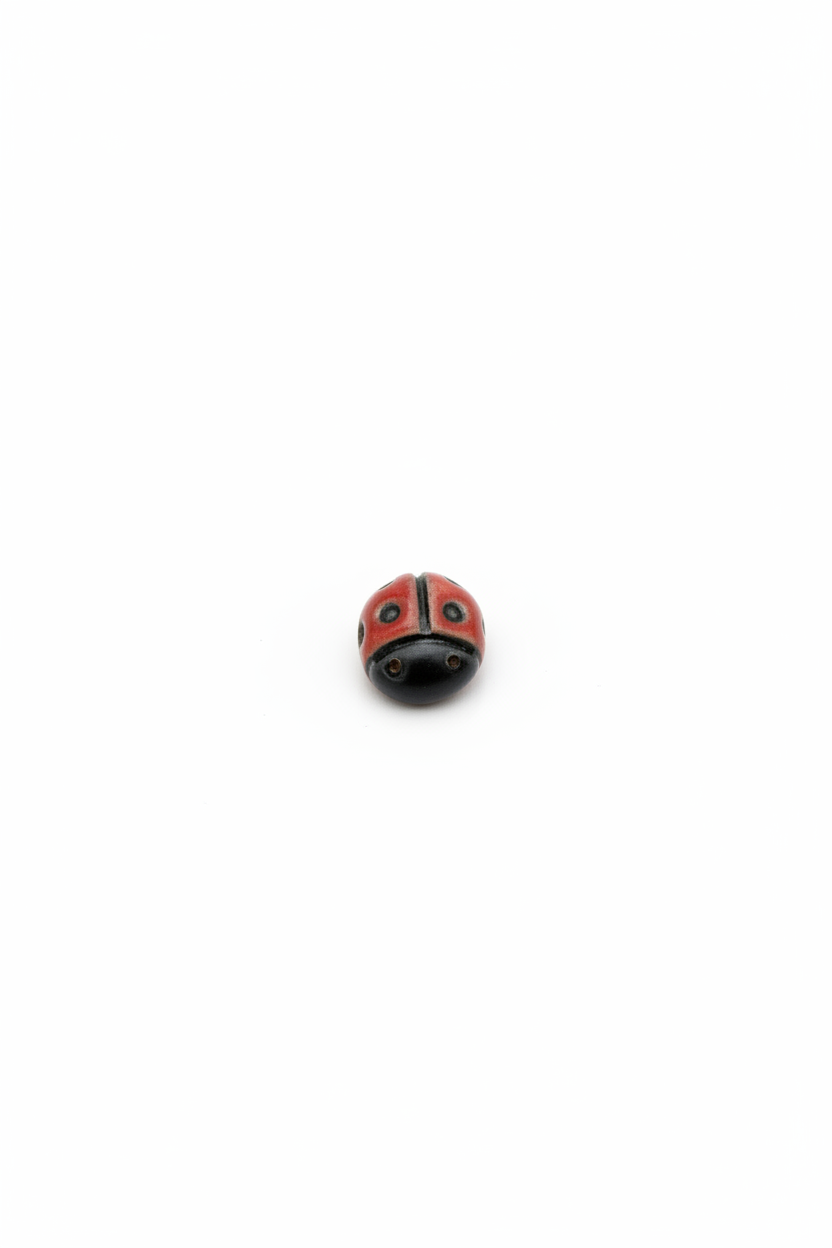 A small round ladybug bead, featuring a red and black painted body with white accents, displayed on a textured off-white fabric background.