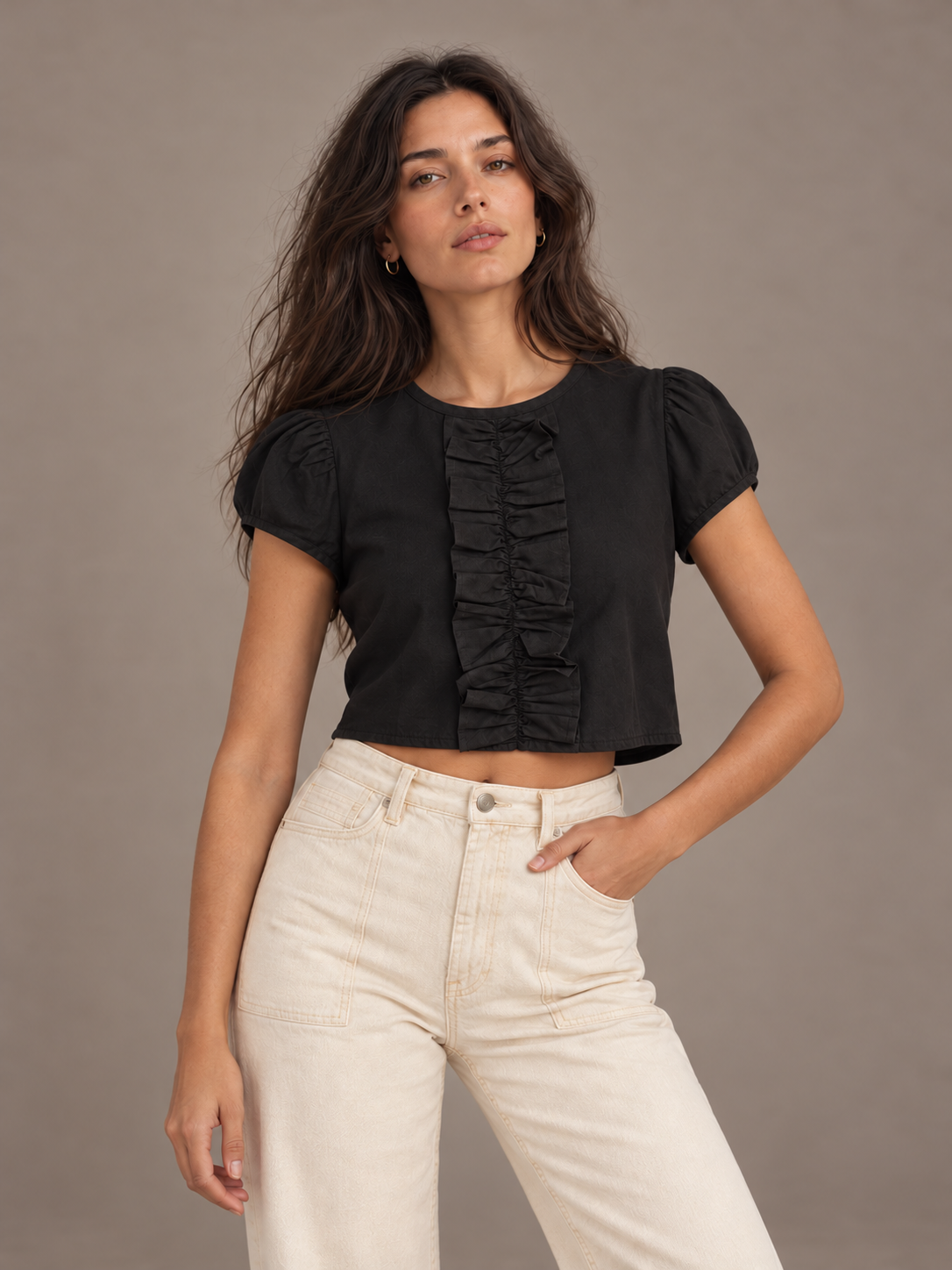 A woman modeling a black short-sleeve crop top with puff sleeves and a vertical ruffle detail down the front, paired with cream-colored pants, against a solid taupe studio background.