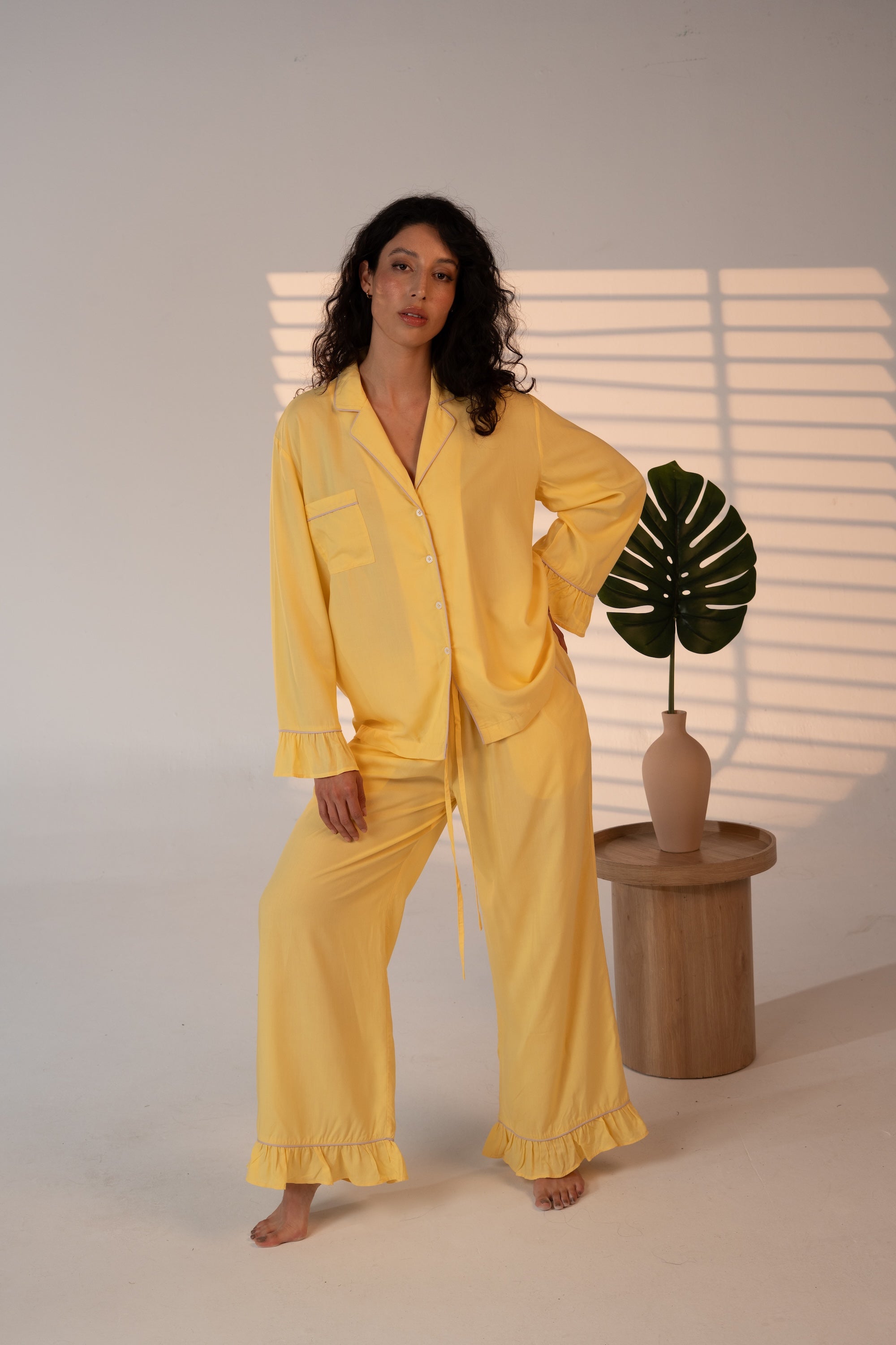 Woman wearing a yellow pajama set standing in a softly lit room with a plant on a small table.