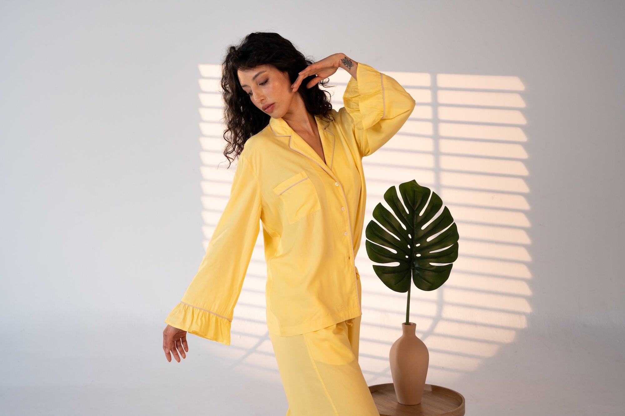 Woman in yellow pajama set standing next to a plant with sunlight filtering through blinds.
