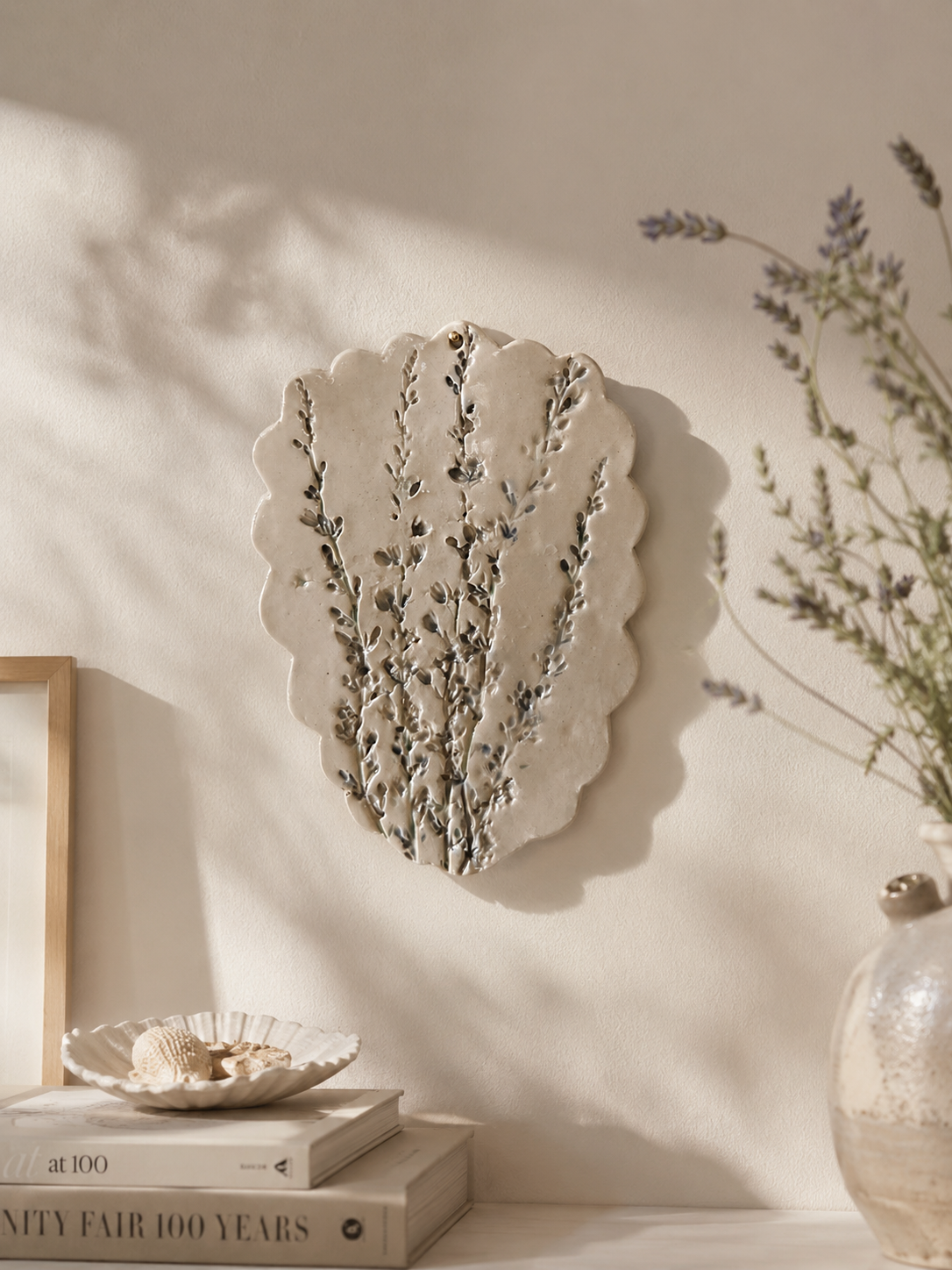 A decorative wall plaque shaped like a scalloped leaf with raised botanical stems, mounted on a warm beige wall. The scene includes soft sunlight shadows, stacked books, a small shell dish, and dried lavender stems in a glass vase.