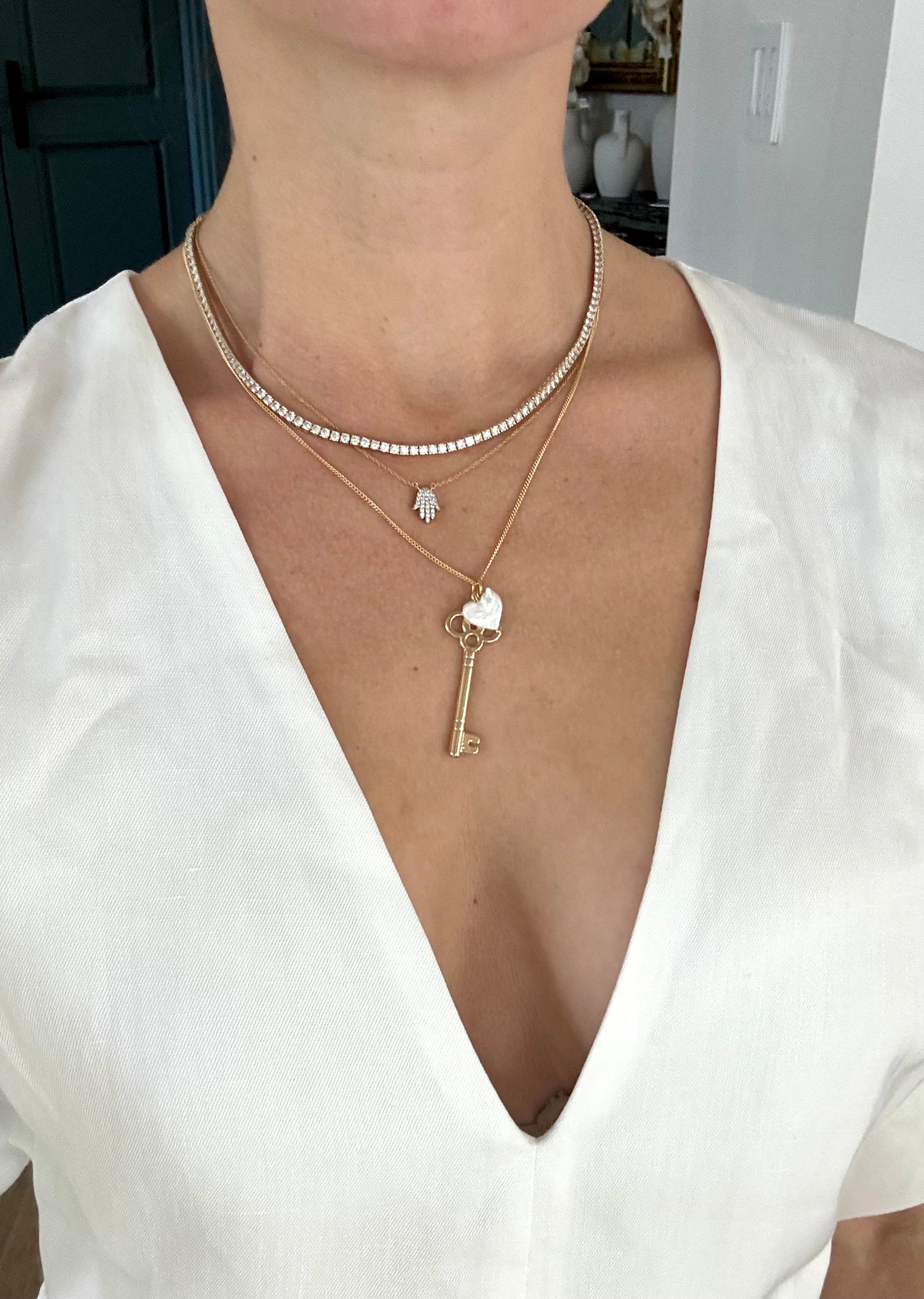 Person wearing a white top with layered necklaces, including a key pendant. Romantic gold key necklace with heart-shaped pearl, part of the Lockheart Theory by Carolina Benoit. A meaningful, affordable luxury gift for her