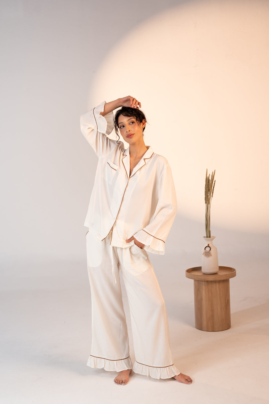 Woman wearing a white pajama set in a minimalistic setting with a wooden side table and decorative plant.