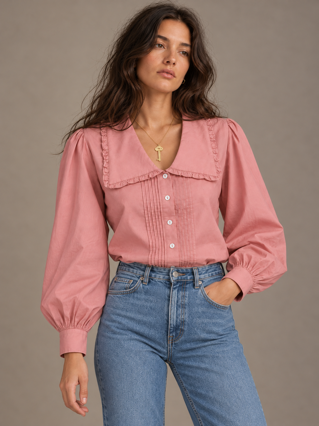 A woman modeling a dusty pink long-sleeve blouse with a large ruffled collar and button front, paired with blue jeans, against a plain taupe background.