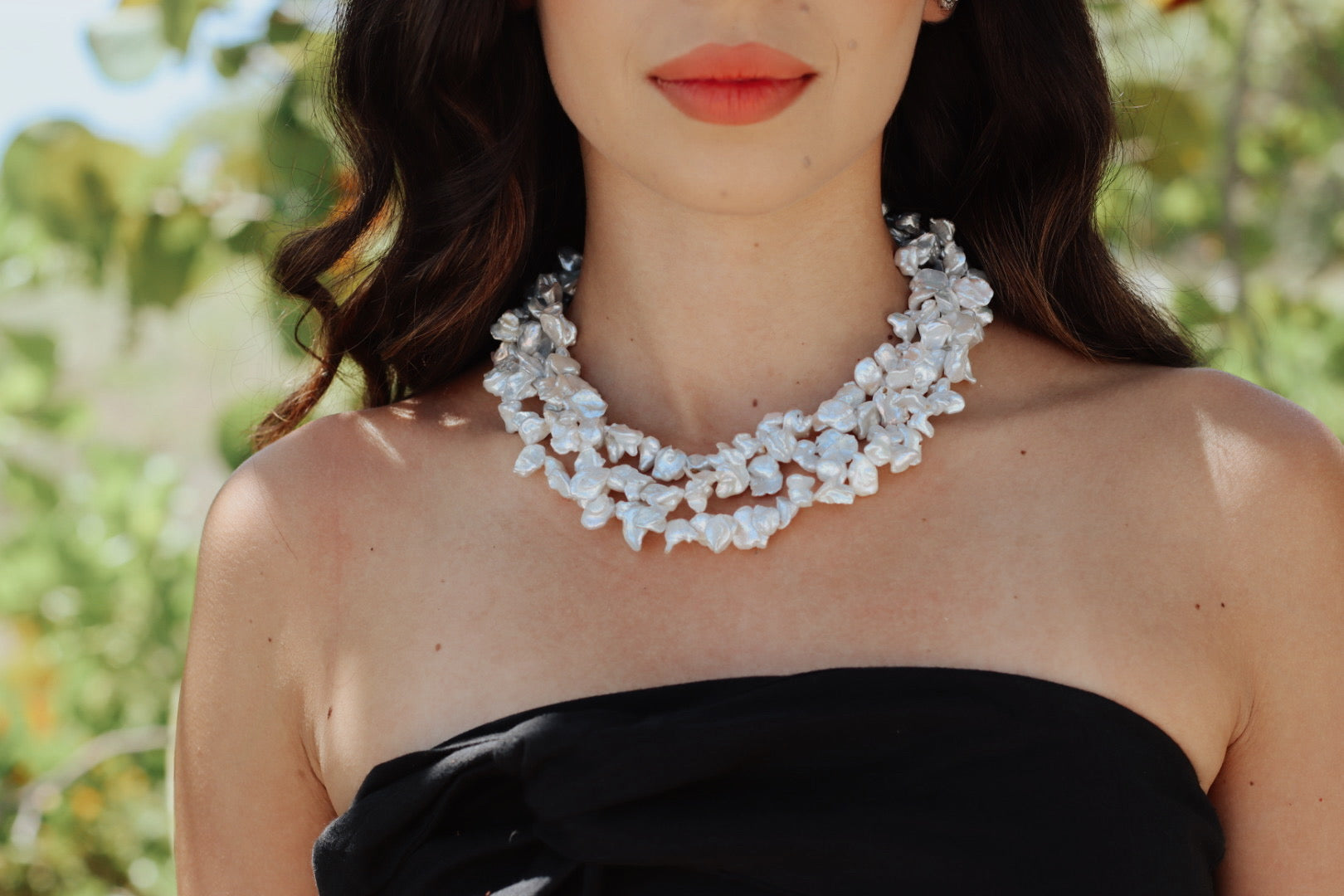 Side view of woman wearing the Baroque Garden Necklace, showing the sculptural pearls layered around her neck, seated among tropical greenery