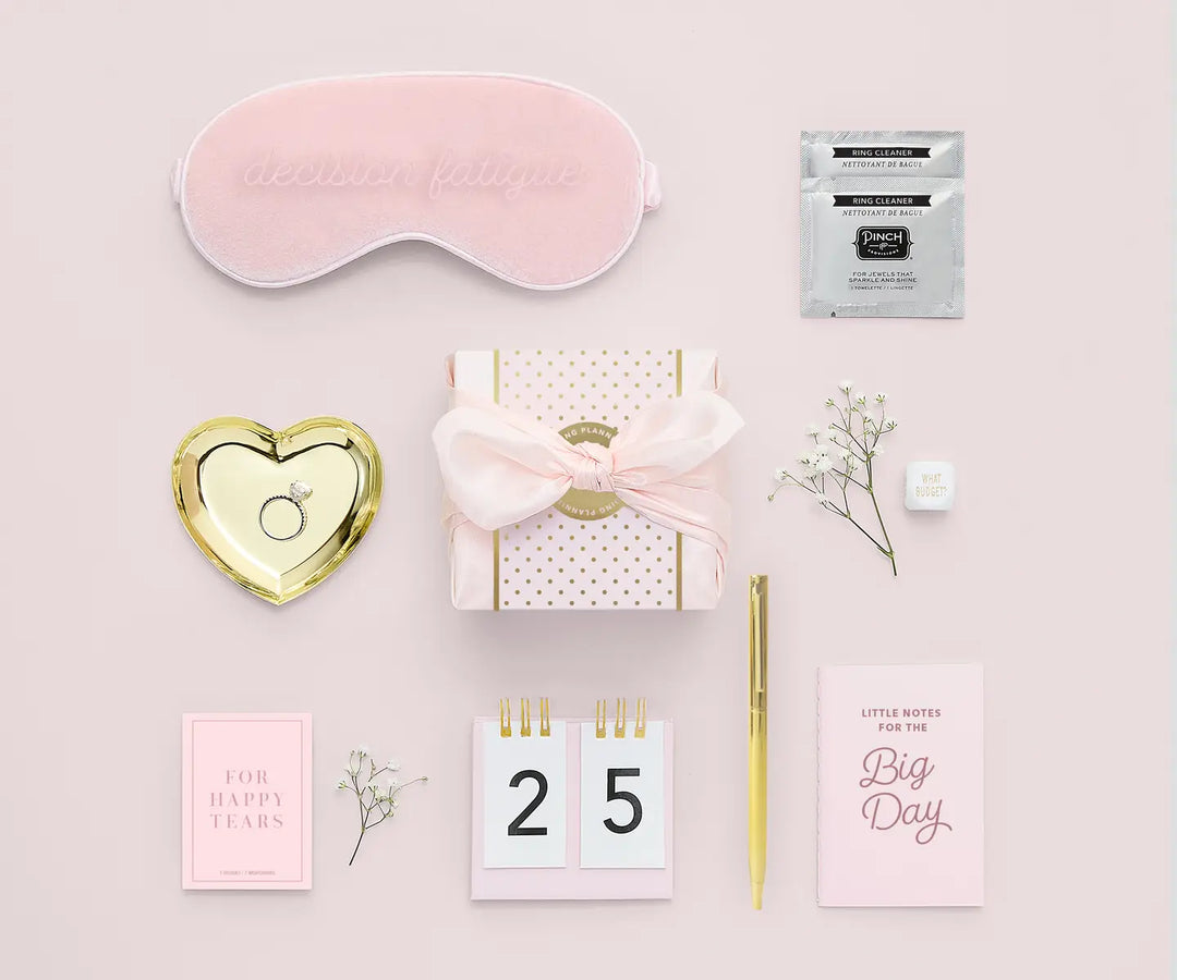 Collection of wedding planning items including a sleep mask, gift box, and stationery on a pink background.