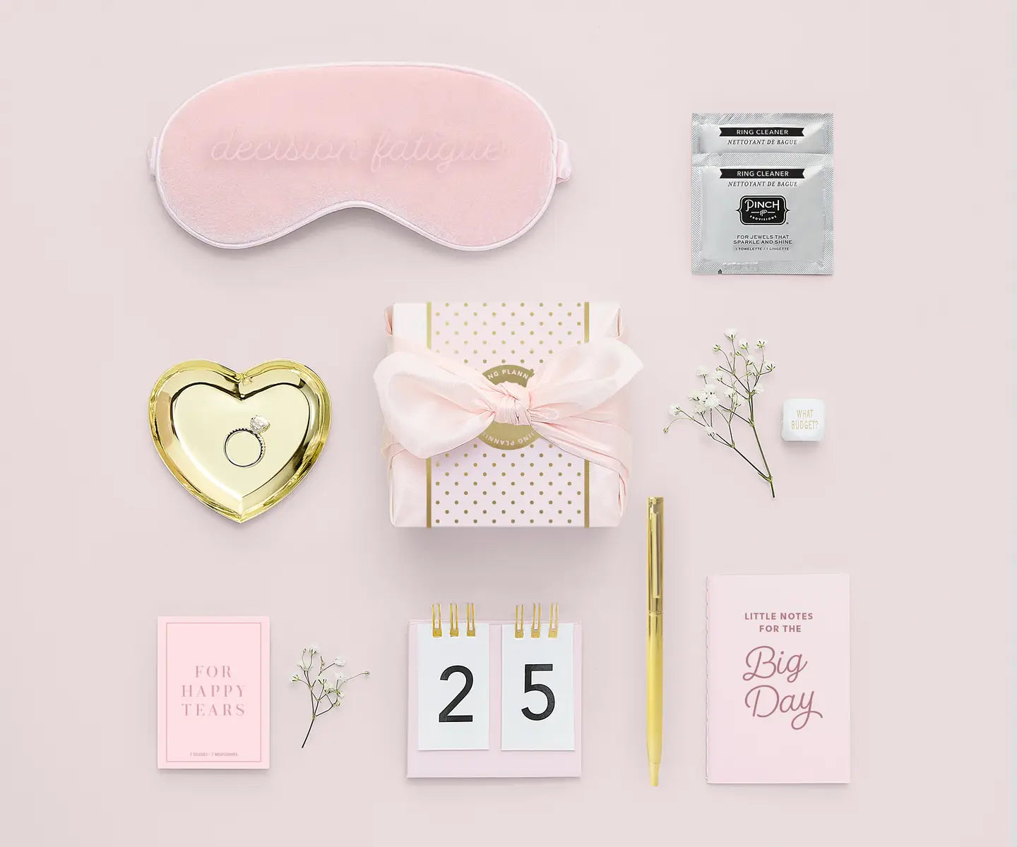 Collection of wedding planning items including a sleep mask, gift box, and stationery on a pink background.