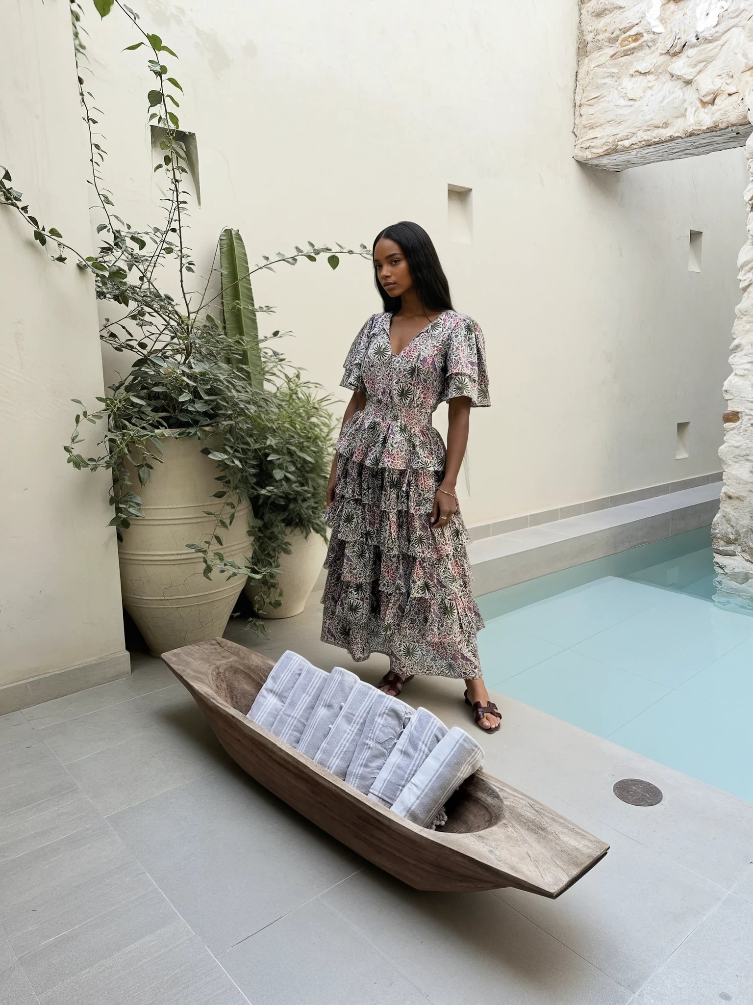 Woman in a floral dress standing next to a wooden boat with towels by a pool. Wearing Carolina Benoit hand block print dress with beautiful tropical background saint remy style. 