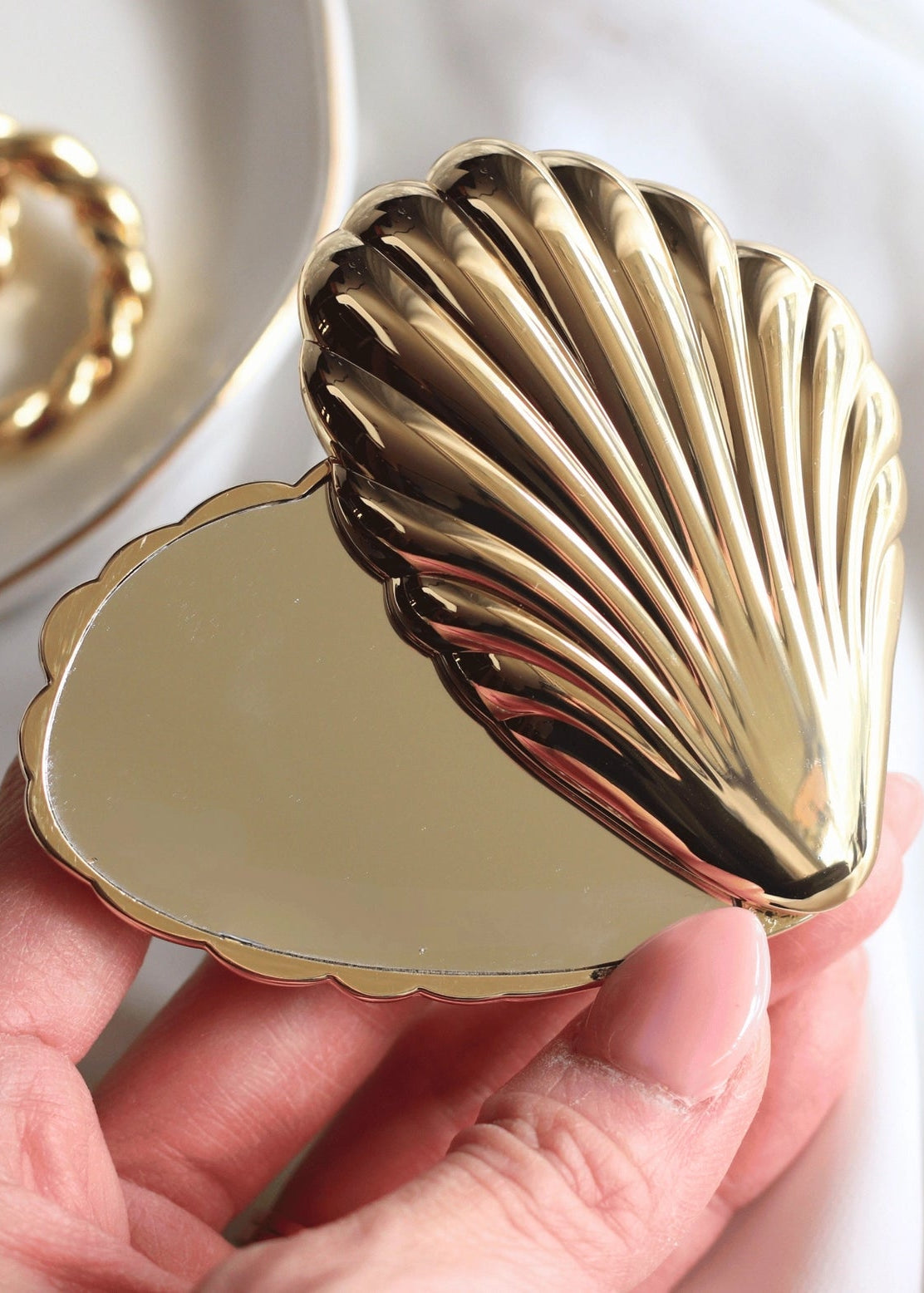Gold shell-shaped compact mirror held by a hand with a blurred background