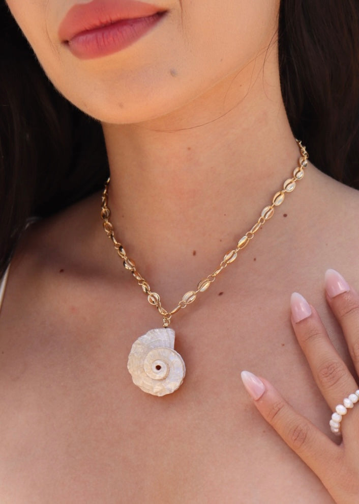 Close-up of a natural spiral shell pendant on a gold-toned cowrie-inspired chain, worn at the collarbone.